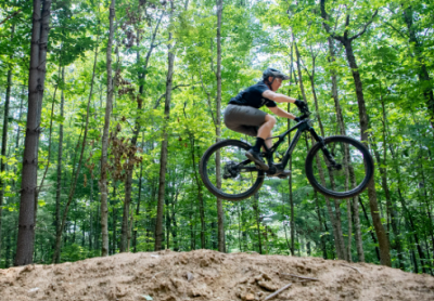 A child rides a mountain bike over dirt jumps in a forested setting