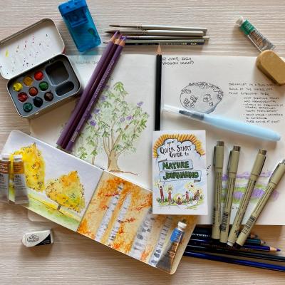 Various art supplies and notebooks arranged on a table with a Nature Journaling guide in the middle.